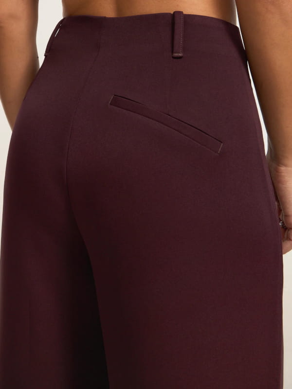 Modelo mostrando detalhes da cintura de uma calça wide leg alfaiatada com pregas duplas bordô.